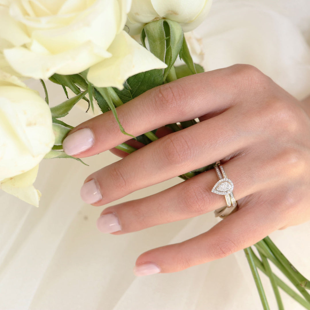 Bride's hand wearing a pear-shaped diamond ring set in yellow gold, holding white roses.